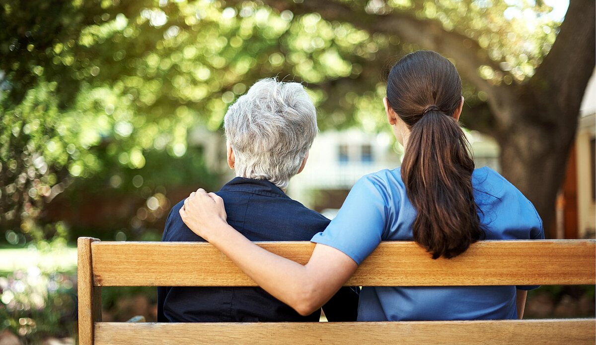Pflegebedürftige Seniorin sitzt mit Pflegerin auf einer Parkbank