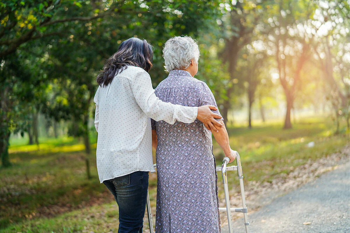 Pflegerin stützt Seniorin mit Rollator beim Spazierengehen im Garten einer Alloheim Senioren-Residenz