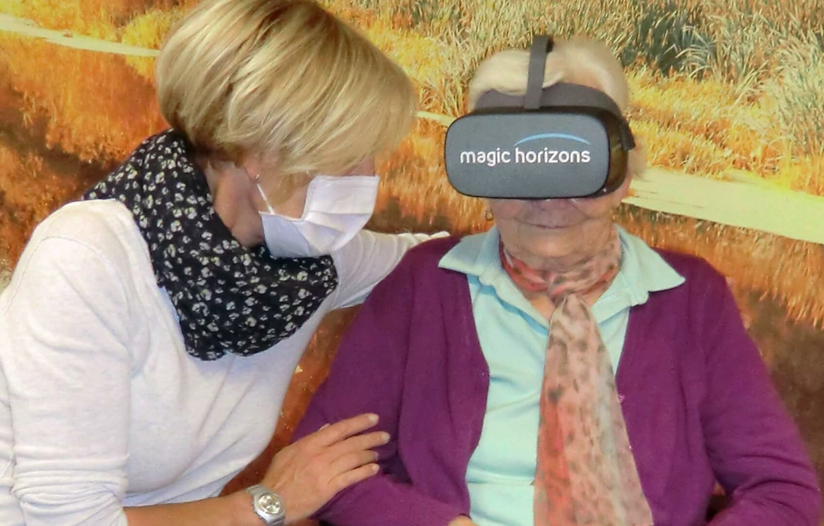Bewohnerin einer Alloheim Senioren-Residenz trägt 3D-Brille, Pflegerin sitzt neben ihr und schaut zu