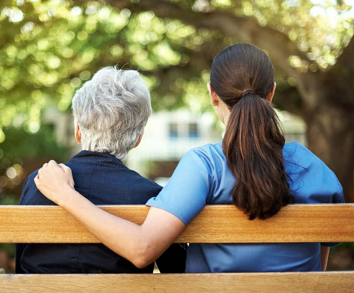 Pflegebedürftige Seniorin sitzt mit Pflegerin auf einer Parkbank
