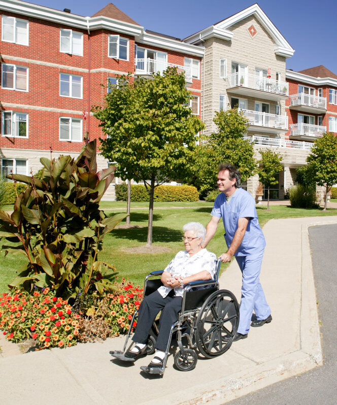 Pflegehilfskraft bei Alloheim fährt Seniorin im Rollstuhl vor einem Pflegeheim