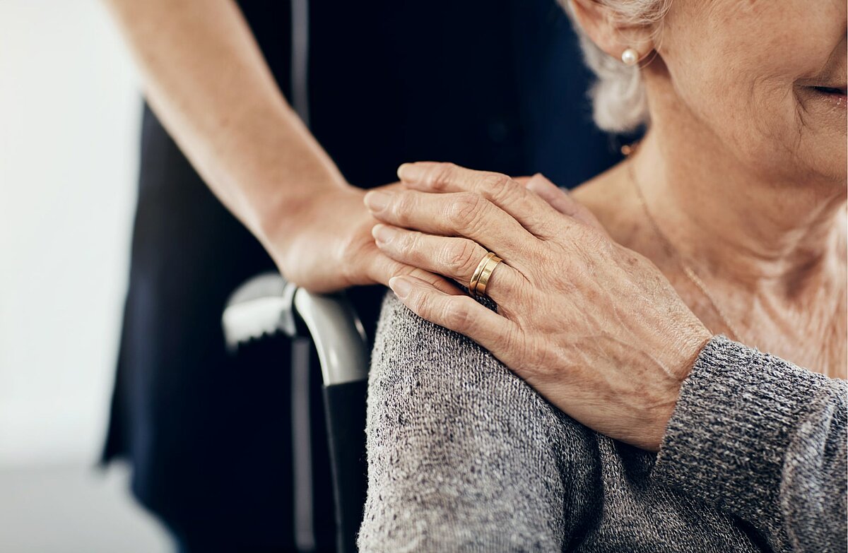 Pflegebedürftige Seniorin im Rollstuhl legt Hand auf Pflegekraft