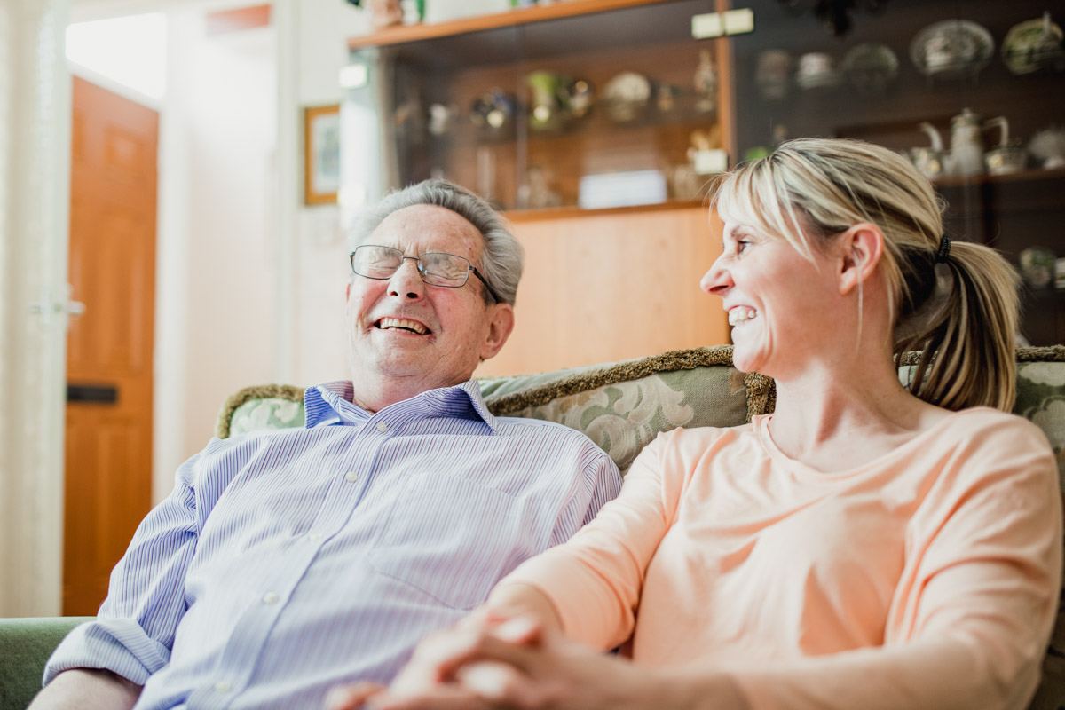 Senior und Pflegefachkraft lachen zusammen auf dem Sofa in einem Alloheim Altenheim