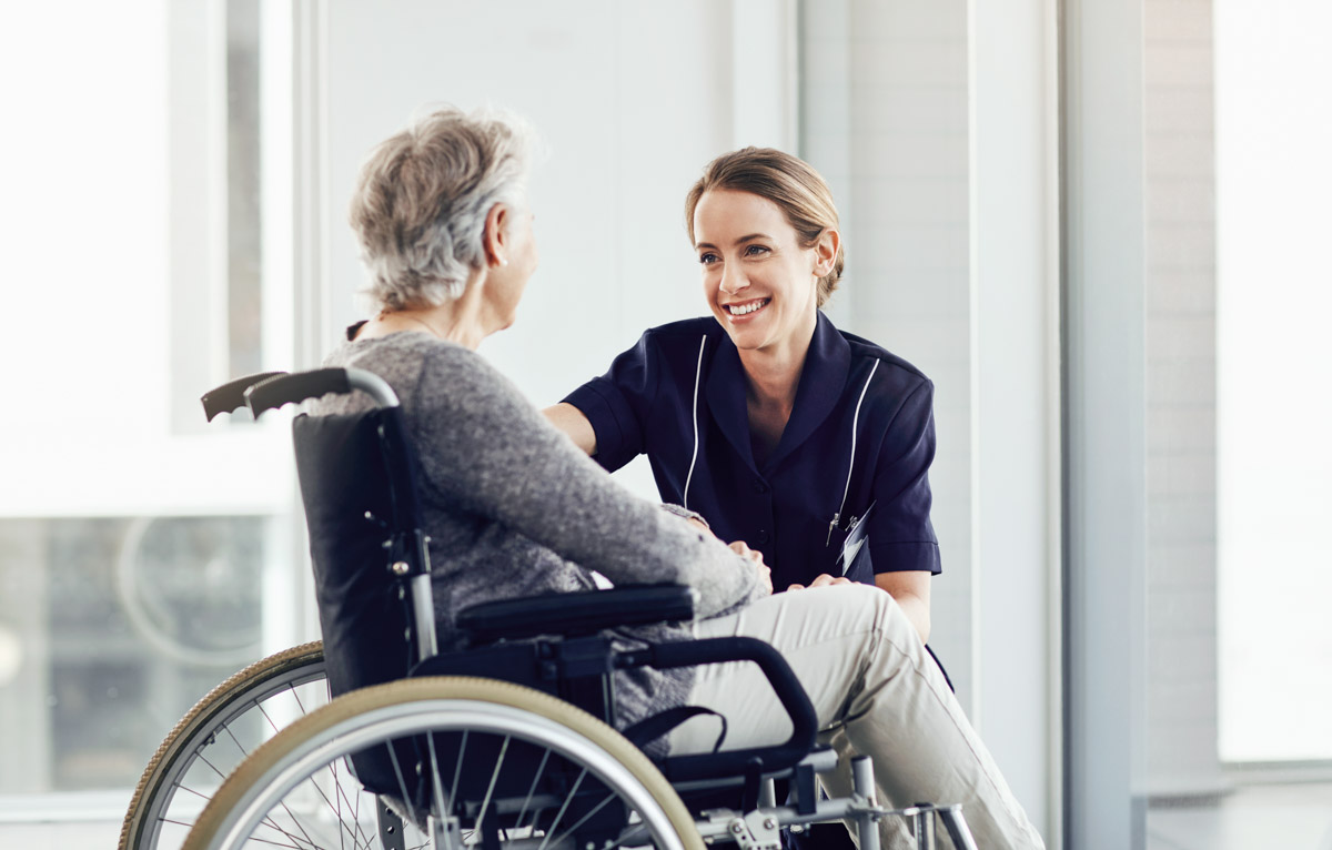 Pflegerin mit Weiterqualifizierung bei Alloheim beugt sich runter zu Seniorin im Rollstuhl