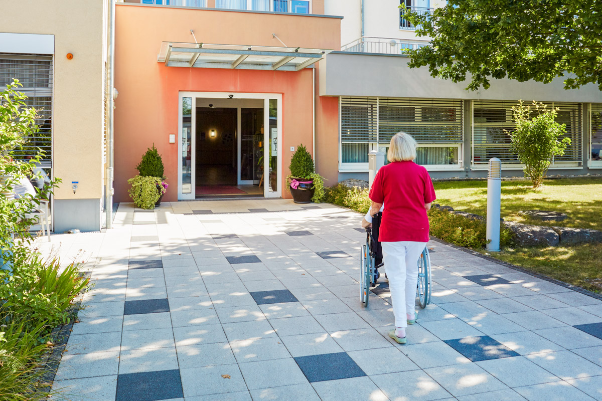 Frau fährt Angehörigen mit Demenz im Rollstuhl in eine Alloheim Senioren-Residenz