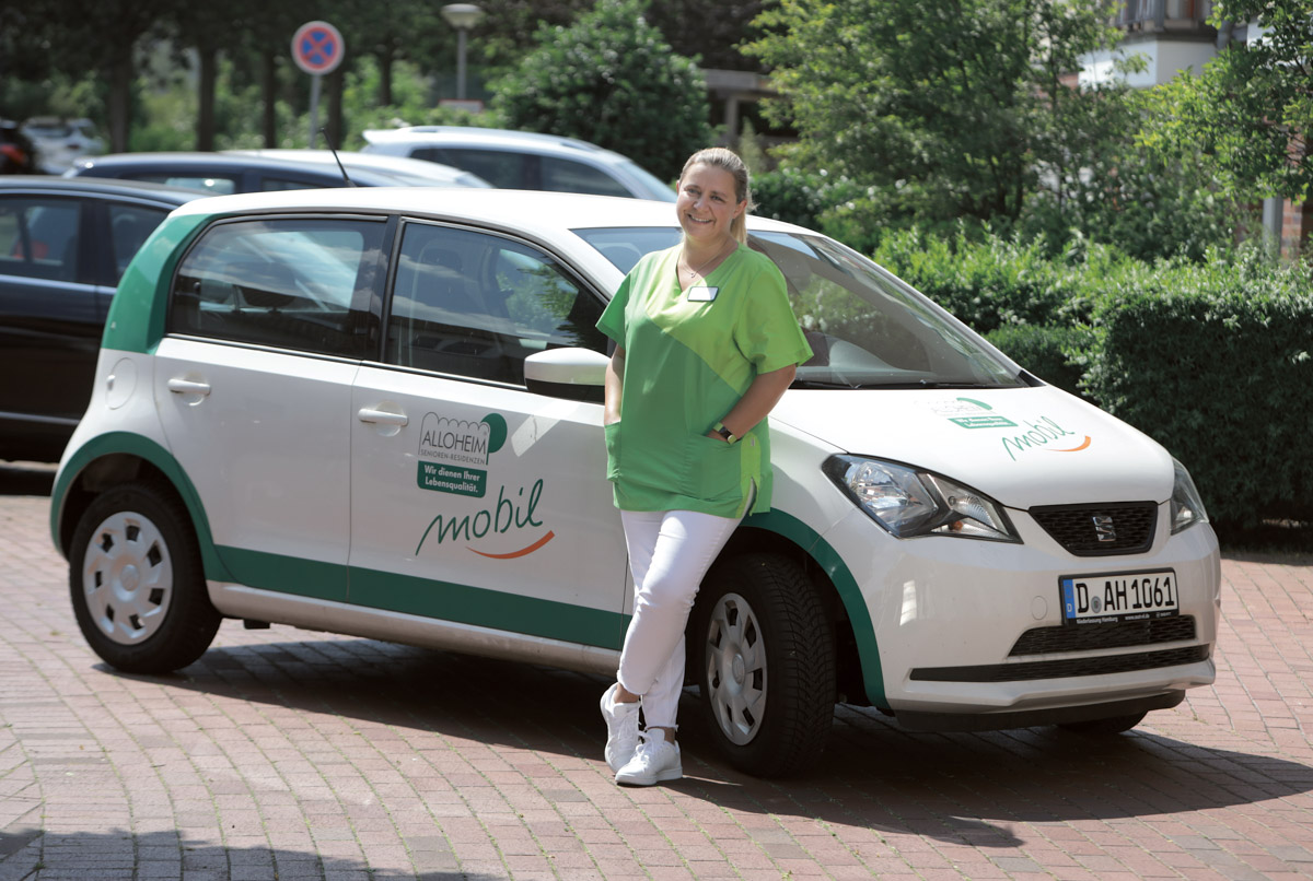 Pflegekraft steht vor dem Auto des ambulanten Pflegediensts Alloheim Mobil