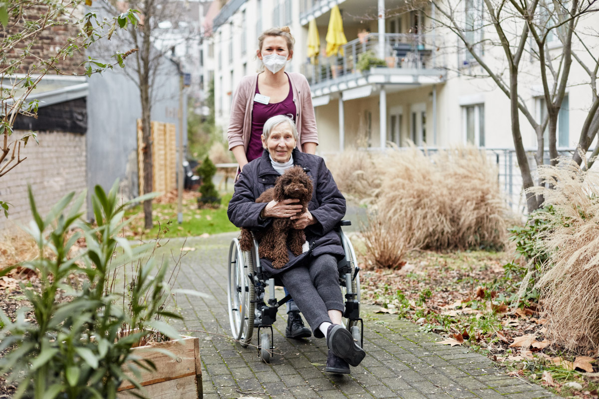 Pflegerin geht mit Bewohnerin der stationären Langzeitpflege bei Alloheim und Hund im Park spazieren