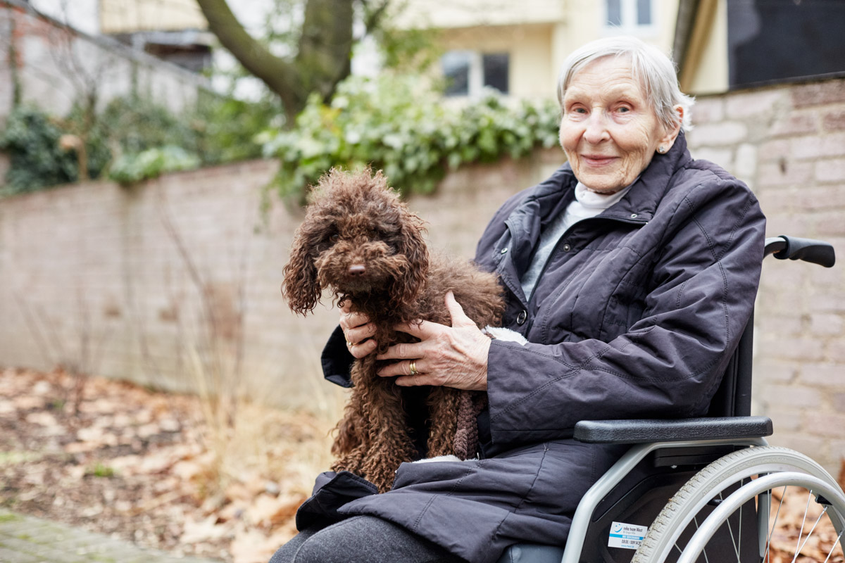 Seniorin im Rollstuhl mit Therapiehund auf dem Schoß im Park einer Alloheim Senioren-Residenz
